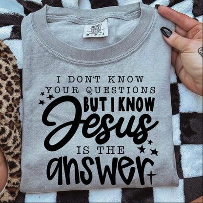 Gray t-shirt with text 'I don't know your questions but I know Jesus is the answer' held by a person.