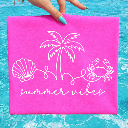 Pink towel with beach-themed designs and 'summer vibes' text held over blue water.
