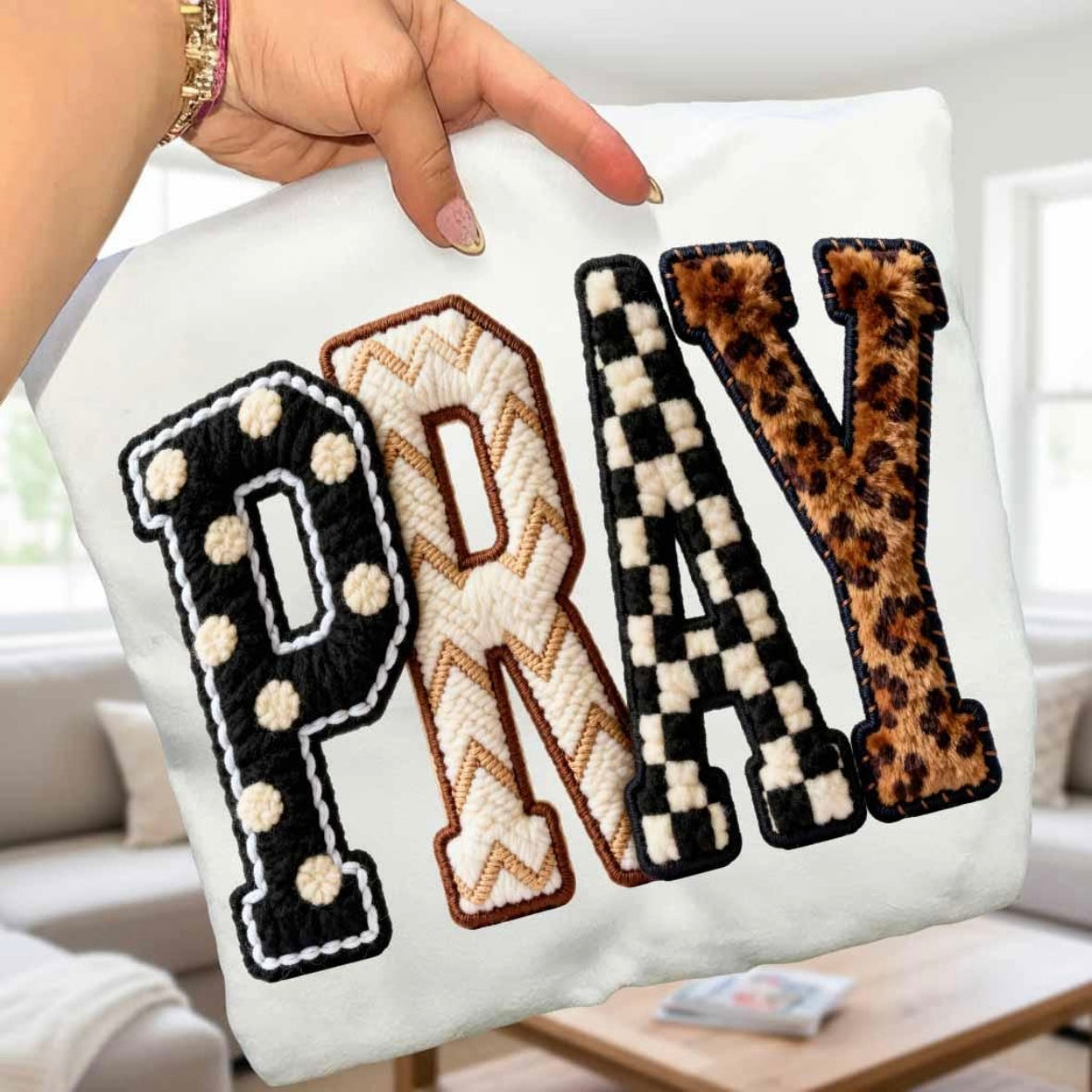White pillow with 'PRAY' letters in various patterns held by a hand.