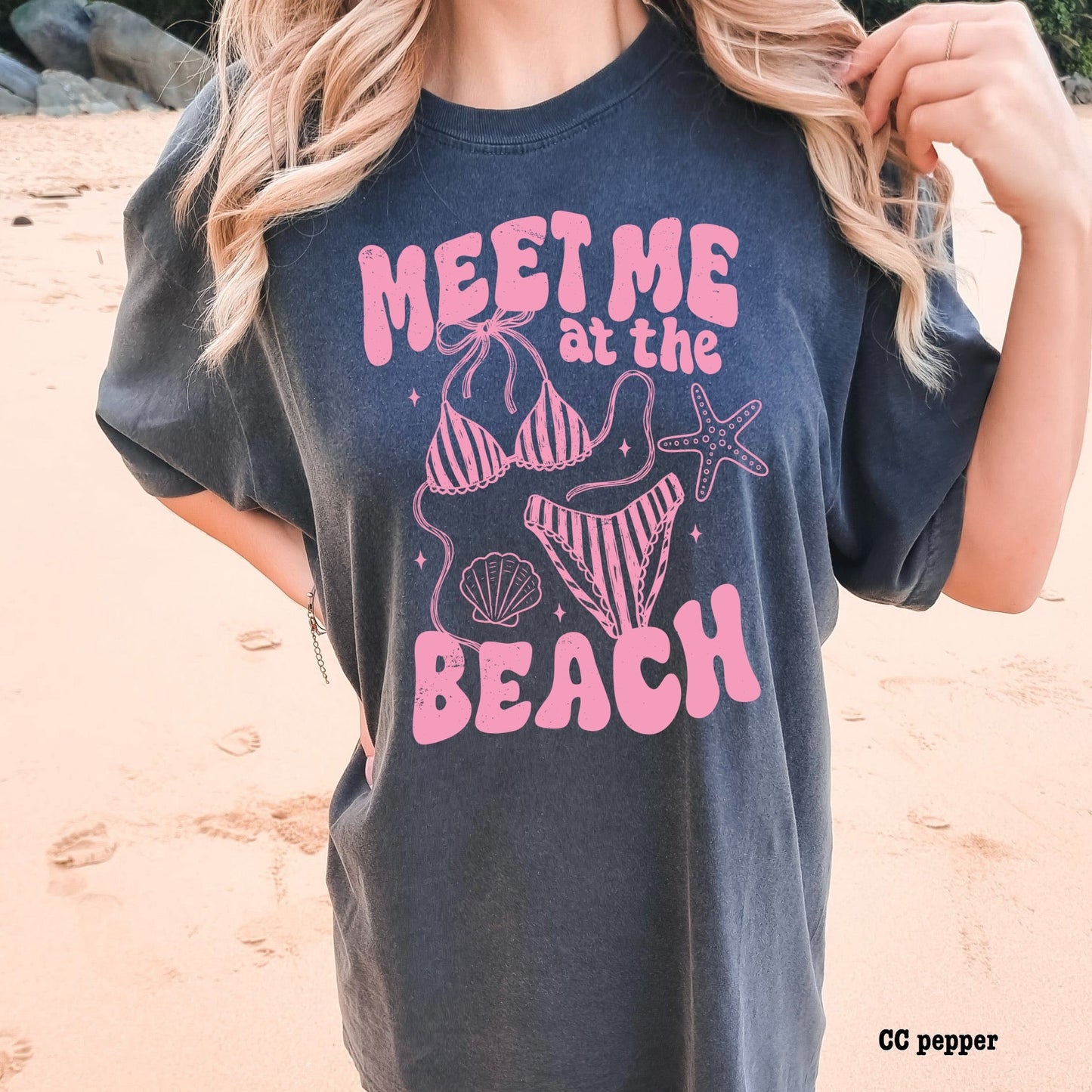 Person wearing a navy t-shirt with pink beach-themed design on a sandy background