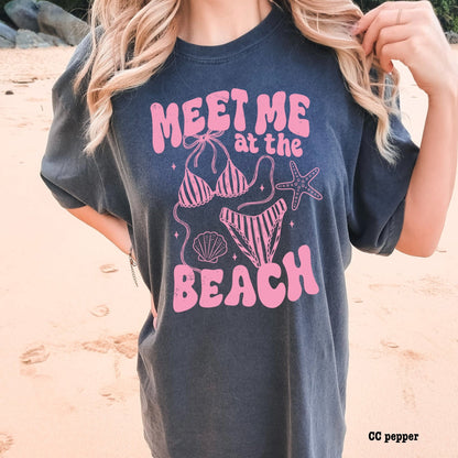 Person wearing a navy t-shirt with pink beach-themed design on a sandy background
