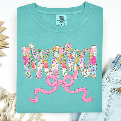 Teal t-shirt with floral and pink text design, placed on a white surface with denim jeans and dried plants.