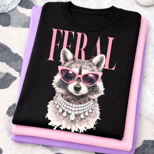 Black t-shirt with a raccoon graphic wearing sunglasses and the word 'Feral' on a pink and purple folded fabric background.