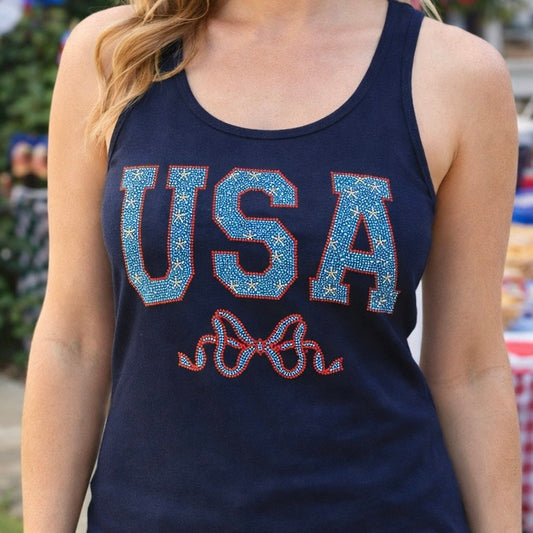 Woman wearing a navy tank top with 'USA' text in blue and red, standing outdoors with American flags in the background.