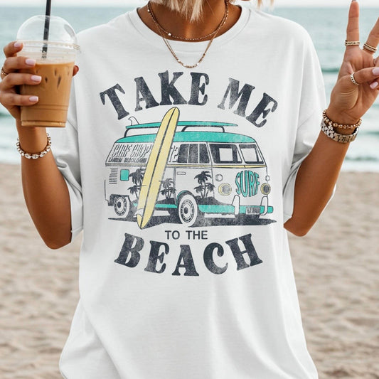 Person wearing a 'Take Me to the Beach' t-shirt with a van and surfboard graphic, holding an iced coffee on a beach.