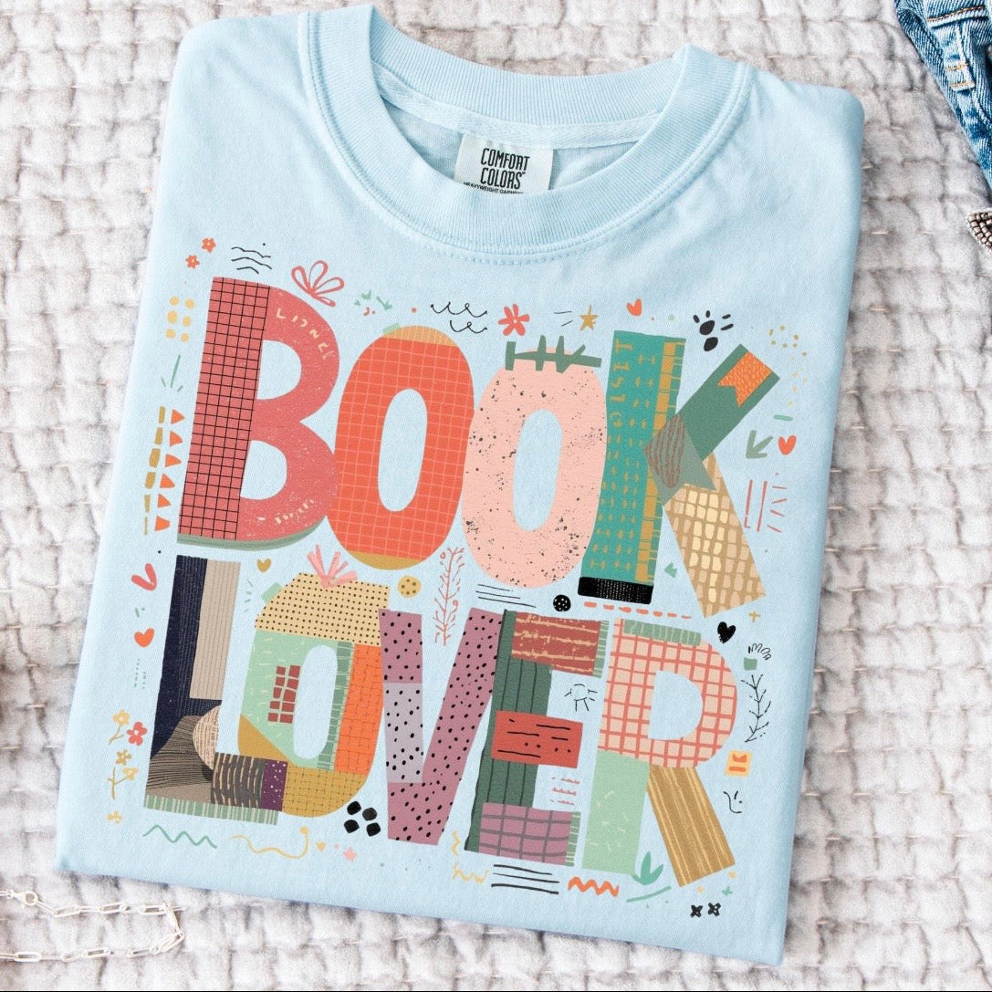 Folded light blue t-shirt with 'BOOK LOVER' text and colorful design on a textured surface.