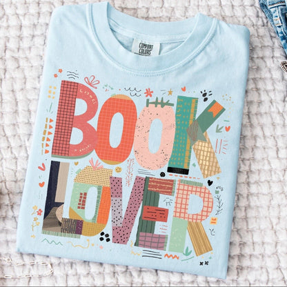 Folded light blue t-shirt with 'BOOK LOVER' text and colorful design on a textured surface.