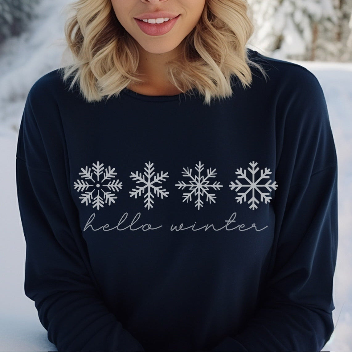 Woman wearing a navy sweater with snowflake designs and 'hello winter' text, standing in a snowy landscape.
