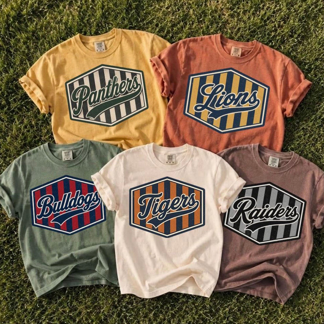 Five graphic t-shirts in different colors (yellow, orange, sage green, white, and mauve) are arranged in a flat-lay on green grass. Each shirt features a large hexagonal graphic with a team name (Panthers, Lions, Bulldogs, Tigers, Raiders) in script over a striped or solid patterned background.