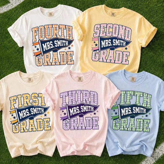Five graphic T-shirts in white, yellow, beige, pink, and light blue are arranged on green turf. Each shirt features a colorful distressed text graphic reading 'MRS.