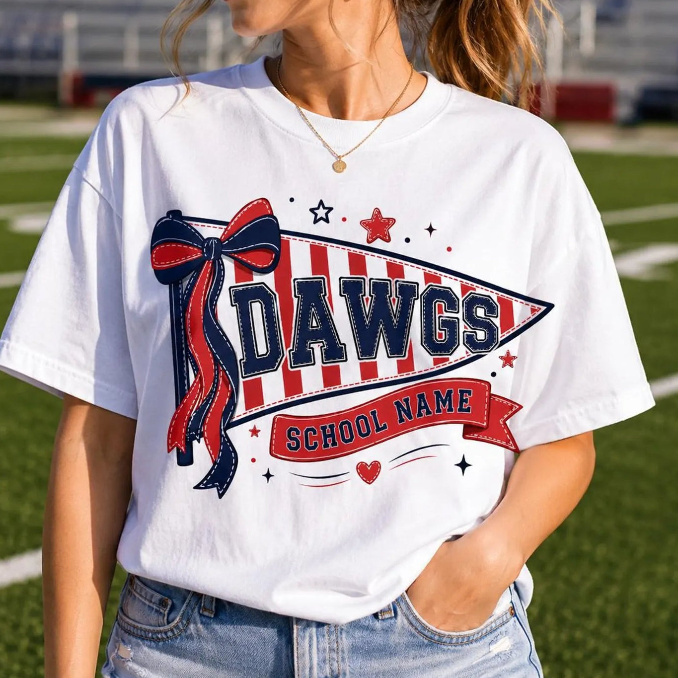A woman wearing a white graphic t-shirt with a red and navy blue 'Dawgs School Name' design on a green football field background.