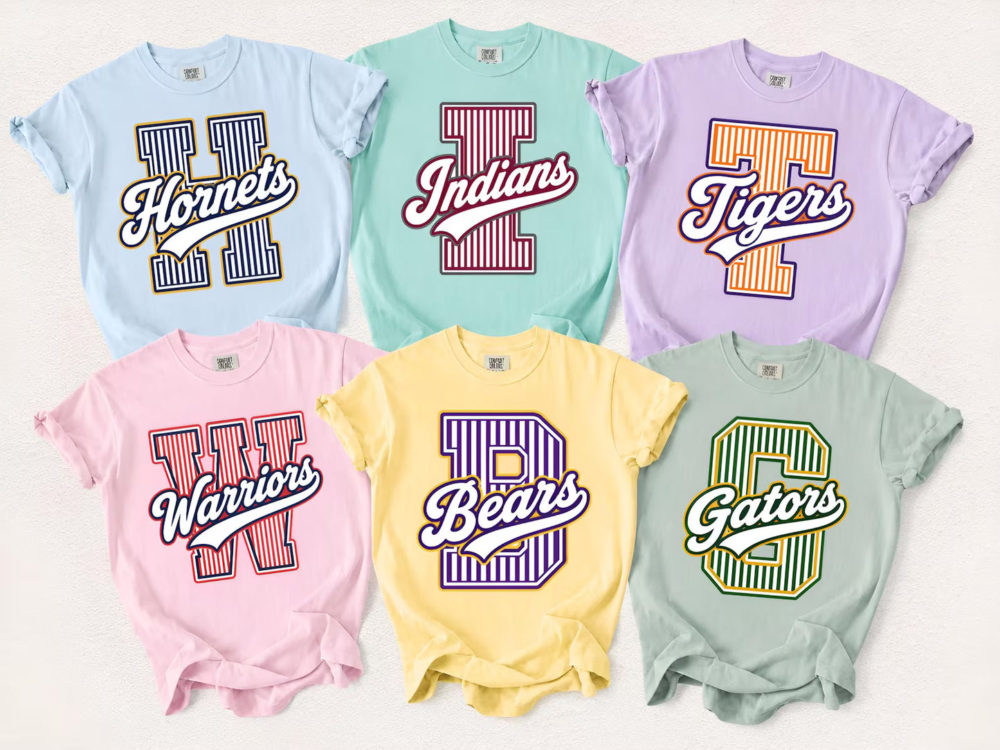 Six short-sleeve graphic T-shirts arranged in two rows on a light neutral background. The top row includes light blue, mint green, and lavender shirts with striped collegiate-style letters reading Hornet, Indians, and Tigers.