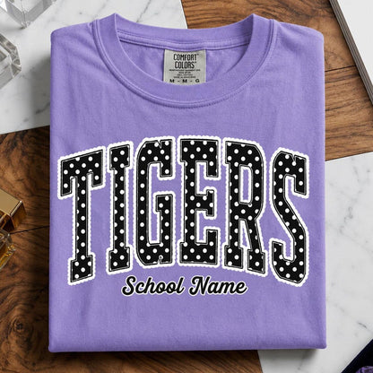 A folded lavender purple t-shirt with 'TIGERS' in black varsity-style letters with white polka dots and a white outline, and 'School Name' printed below, displayed on a wooden surface with marble and crystal props.